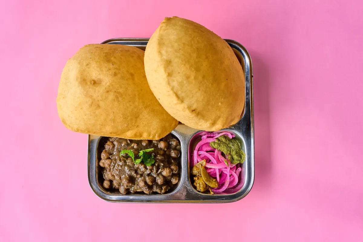 Puffed bhatura breads above a steel tray of chickpeas, sliced pink onion and pickle on a pink surface.
