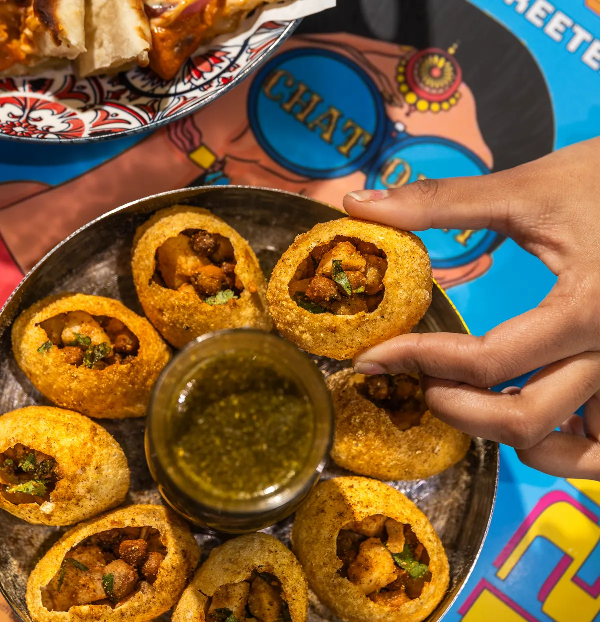 A hand lifts a filled golgappa shell above a plate of shells and a cup of green flavoured water.