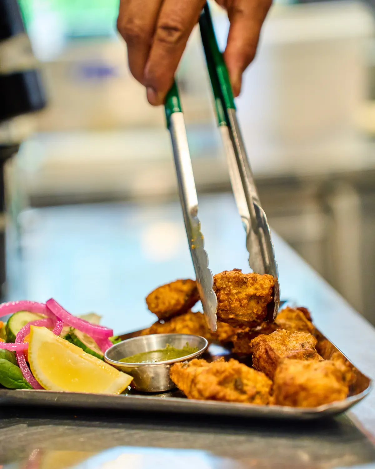 A hand using tongs to arrange golden battered pieces beside pink onion, lemon and chutney.