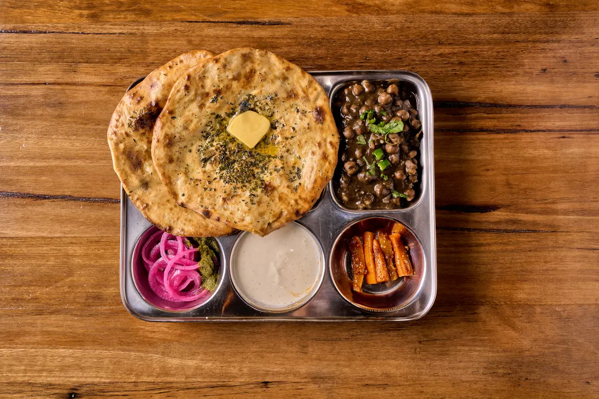 Butter-topped kulcha with chickpeas, pink onions, yoghurt and pickle in a divided steel tray.