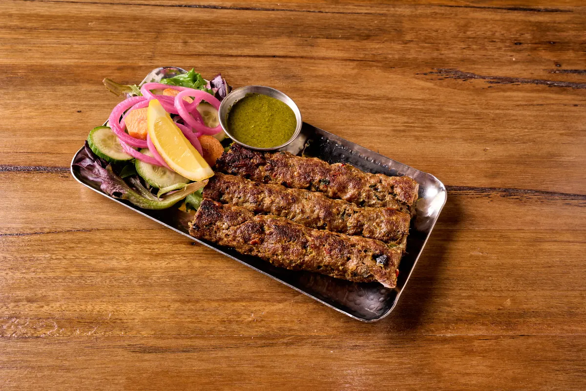 Grilled seekh kebabs beside lemon, pink onion and green chutney on a steel plate.