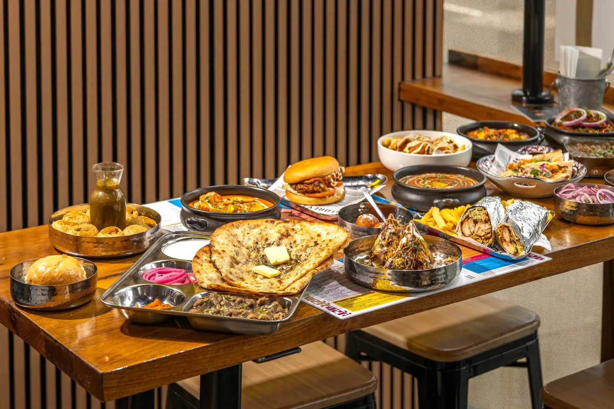 A spread of curries, a burger, naan tacos and wraps on a wooden restaurant table.